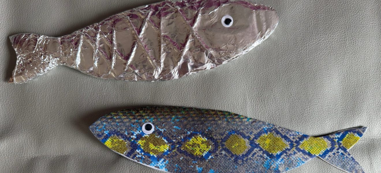 Zwei Fische, die aus unterschiedlichen Materialien gebastelt wurden. Einer der Fische ist silberfarben, der andere weist ein blau-gelbes Muster auf.