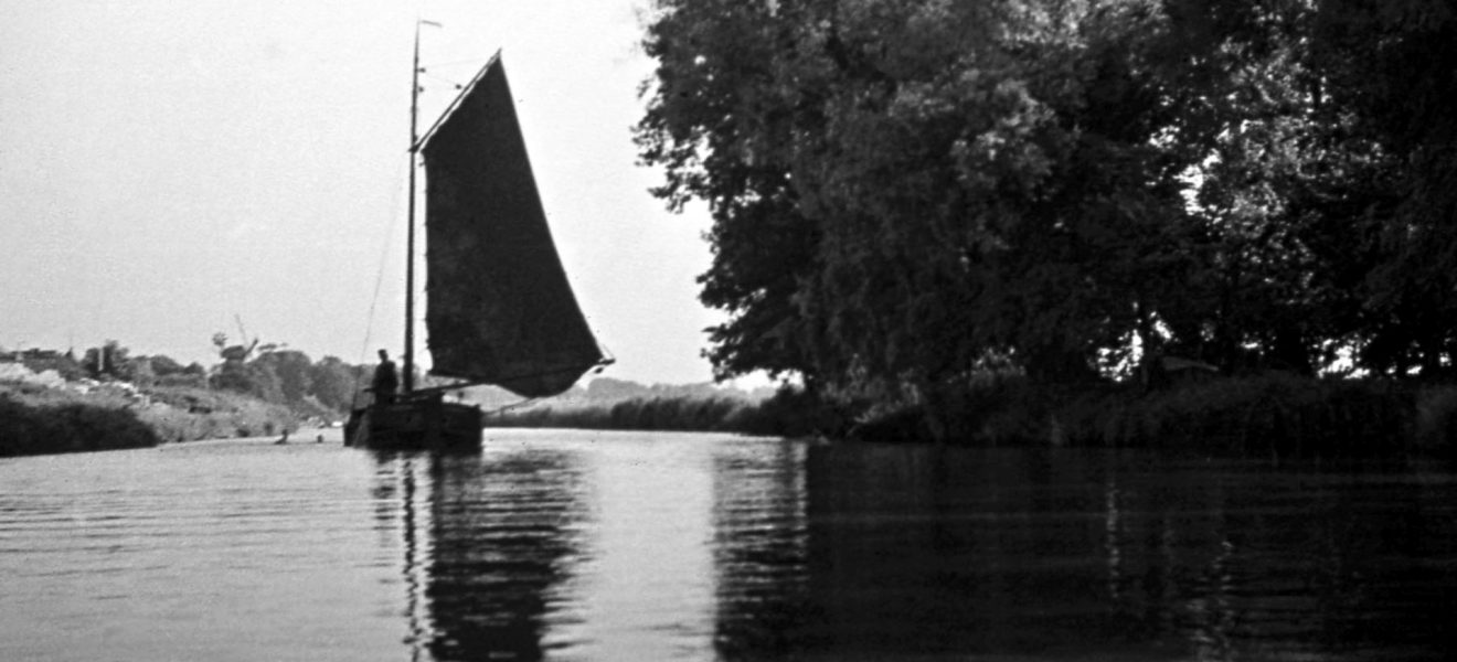 Schwarz-weiß Fotografie eines Torfschiffes auf dem Wasser. Am rechten Ufer stehen große Bäume