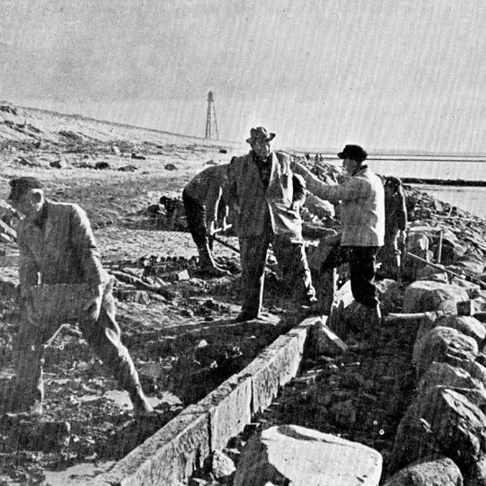 Deichbauarbeiten zwischen Upleward und Campen, mit Blick in Richtung des Campener Leuchtturms, o. J. (Archiv des Entwässerungsverbands der Deichacht Krummhörn) Schwarz-weiß Fotografie mit Arbeitern an einem Deich